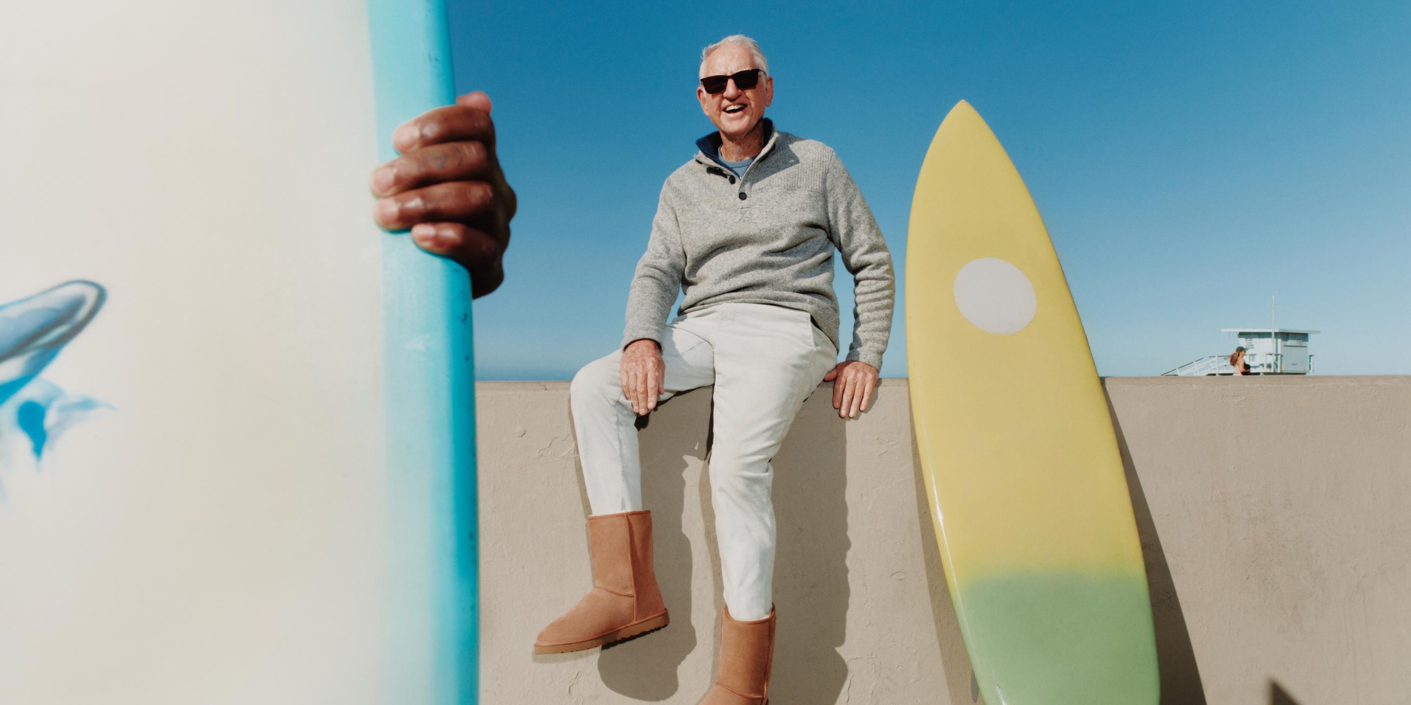 A person sitting on a fence next to a surfing board, wearing sunglasses and Classic Short II boots in chestnut. 