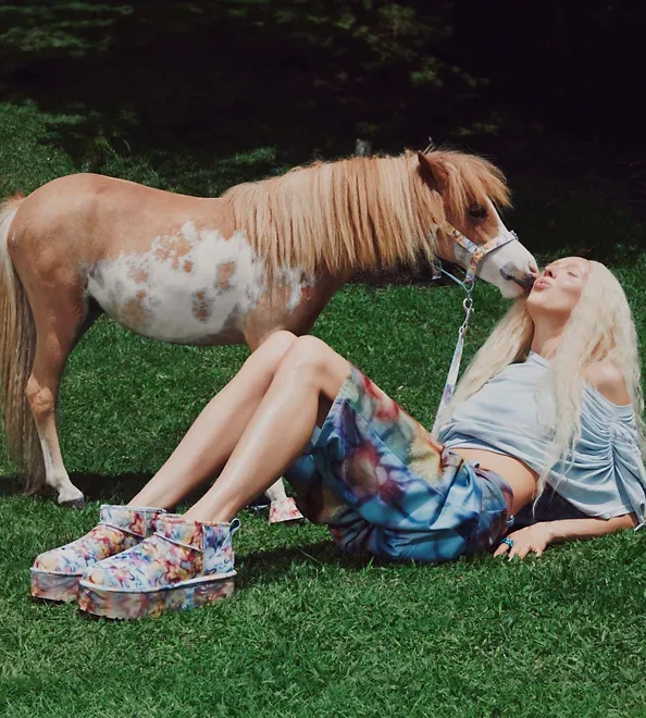 A woman wearing UGG Collina Ultra Mini Platform is lying on the grass while a pony kisses her face.