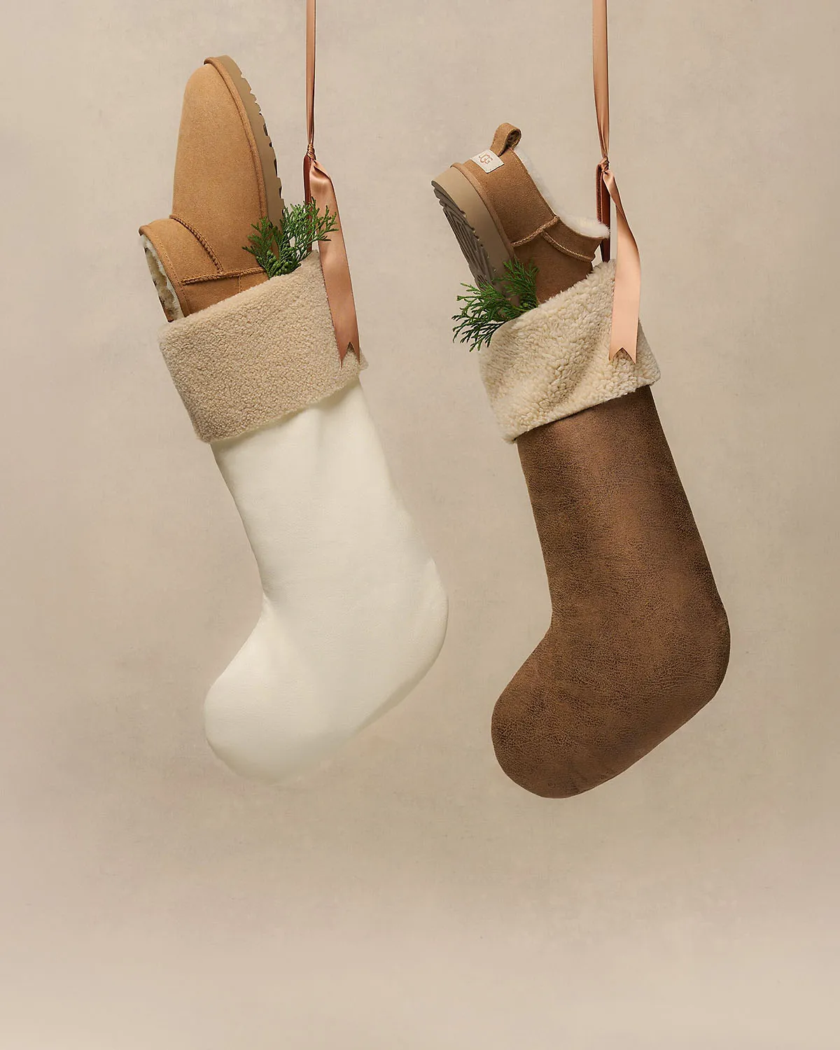 Two slippers hang from pink ribbons inside cream and brown stockings against a neutral wall.