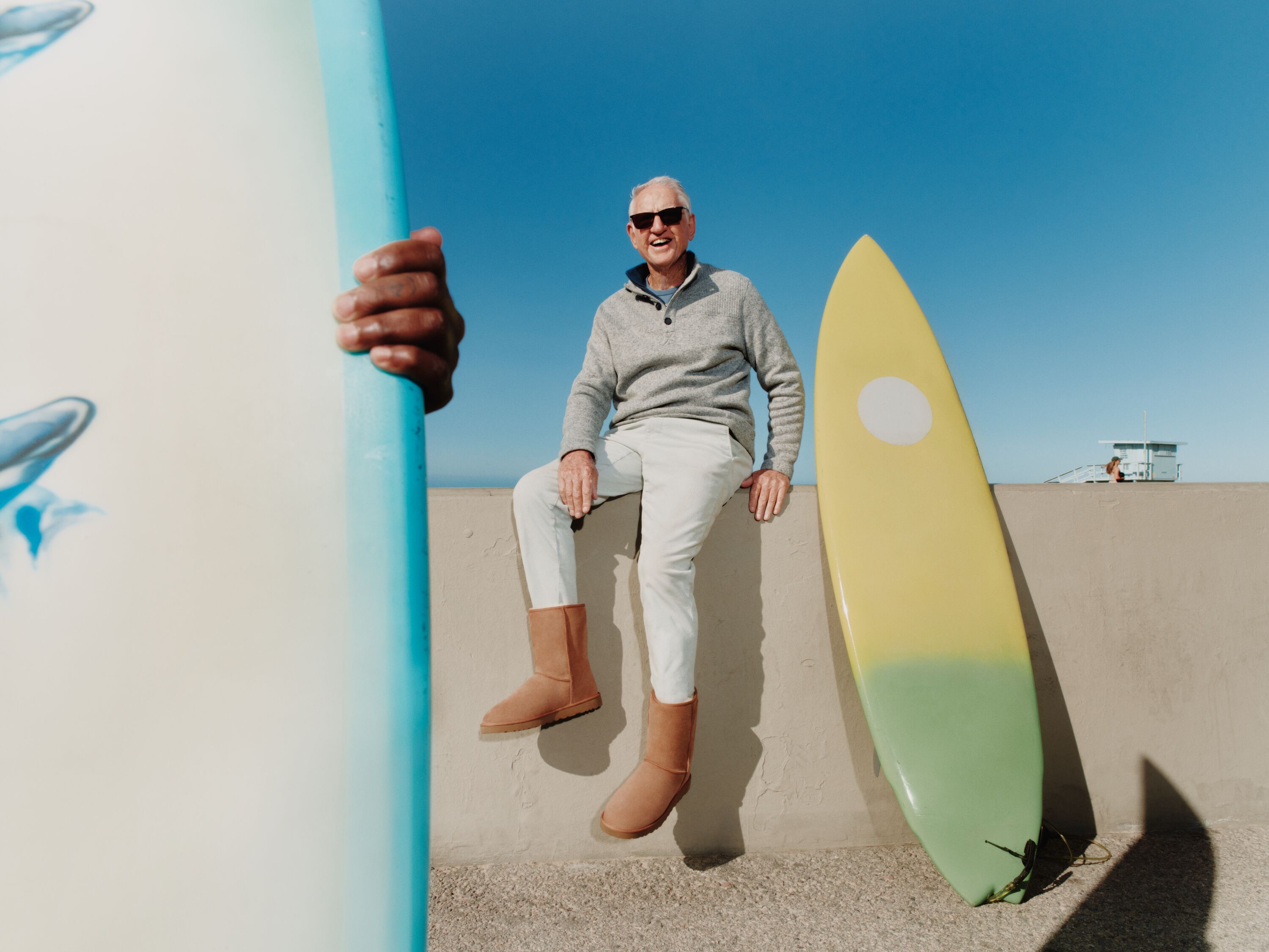 Découvrez notre histoire Le fondateur d’UGG, Brian Smith, assis sur un rebord de plage portant les bottes courtes UGG Classic pour hommes de couleur marron.