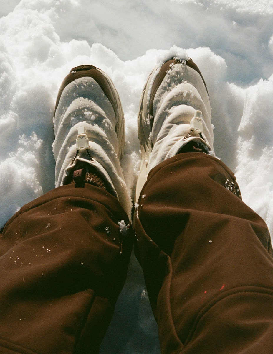 Gros plan des jambes d’une personne posées sur la neige et portant des chaussures HOKA Kaha 2 Frost Moc GTX Halfdays.