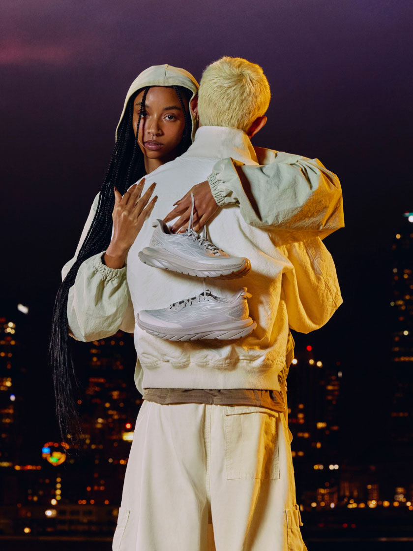 Trois personnes s’embrassant, la ville en arrière-plan tout en tenant des chaussures HOKA Mach Remastered.