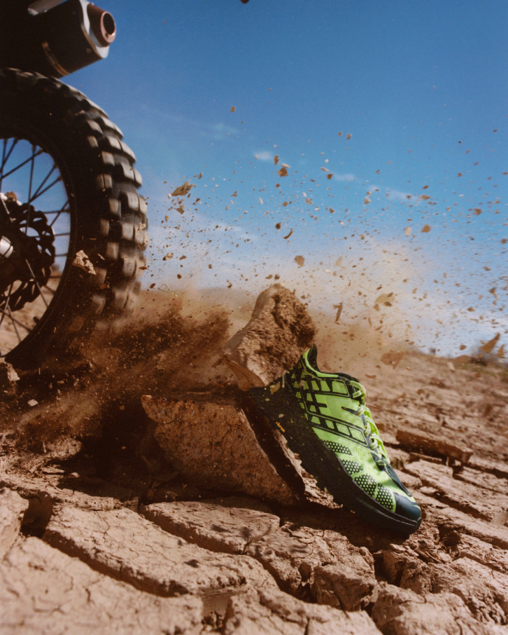Chaussure HOKA Speedgoat 2 vert néon avec de la terre soulevée par un pneu de moto.