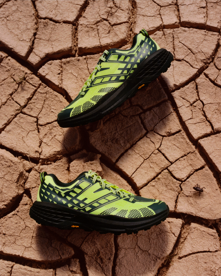 Photo aérienne de baskets HOKA Speedgoat 2 vert néon et noires sur une surface desséchée et craquelée de boue, avec un petit scarabée à proximité.