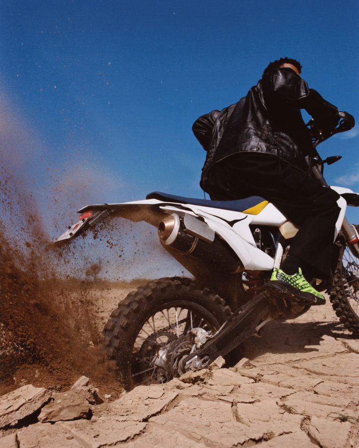 Un pilote portant des chaussures HOKA Speedgoat 2 vert néon accélère une moto tout-terrain blanche sur une terre desséchée.
