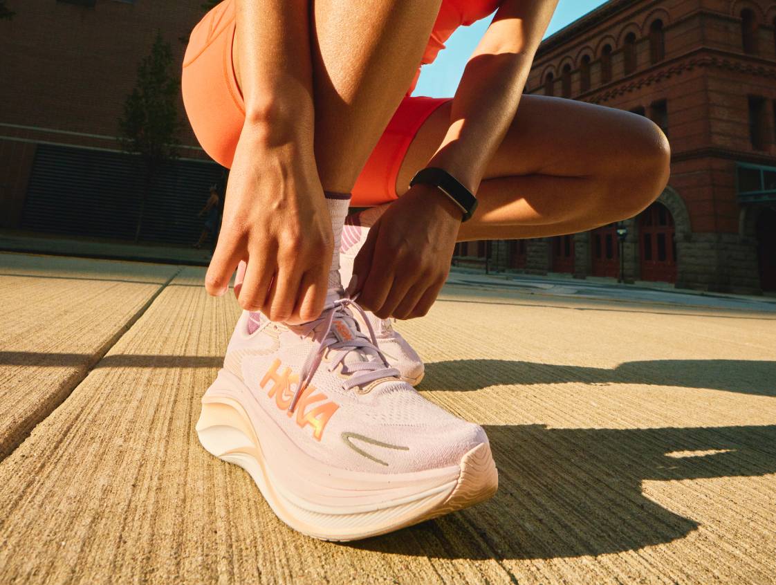 Gros plan d’un coureur en train de nouer les lacets d’une basket HOKA rose pâle dans une rue ensoleillée de la ville.