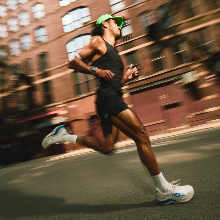 Vue latérale d’un coureur dans une rue de la ville montrant des chaussures de course HOKA blanches et bleues.
