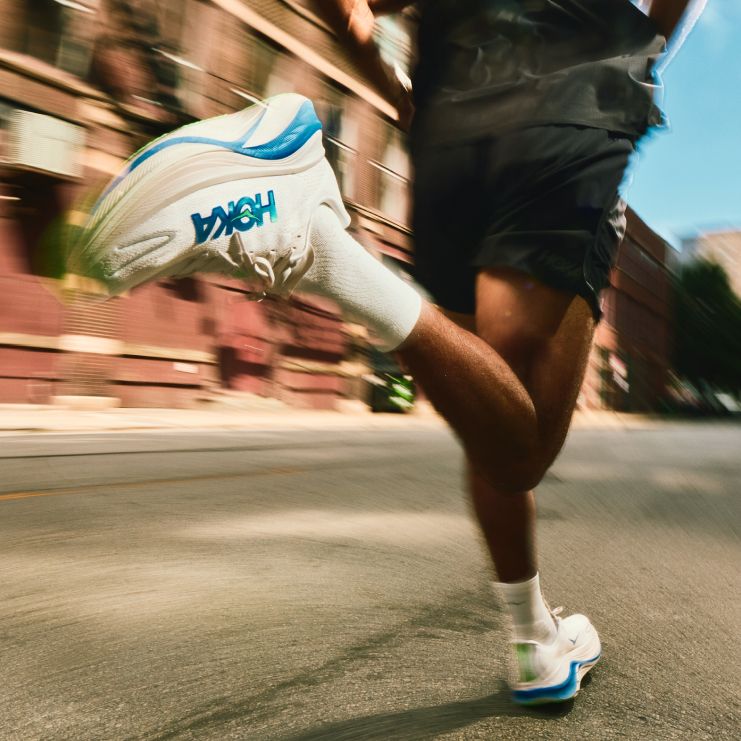 Photo d’action en pleine foulée se concentrant sur des baskets de course HOKA à semelles épaisses, blanches et bleues, avec une marque visible.