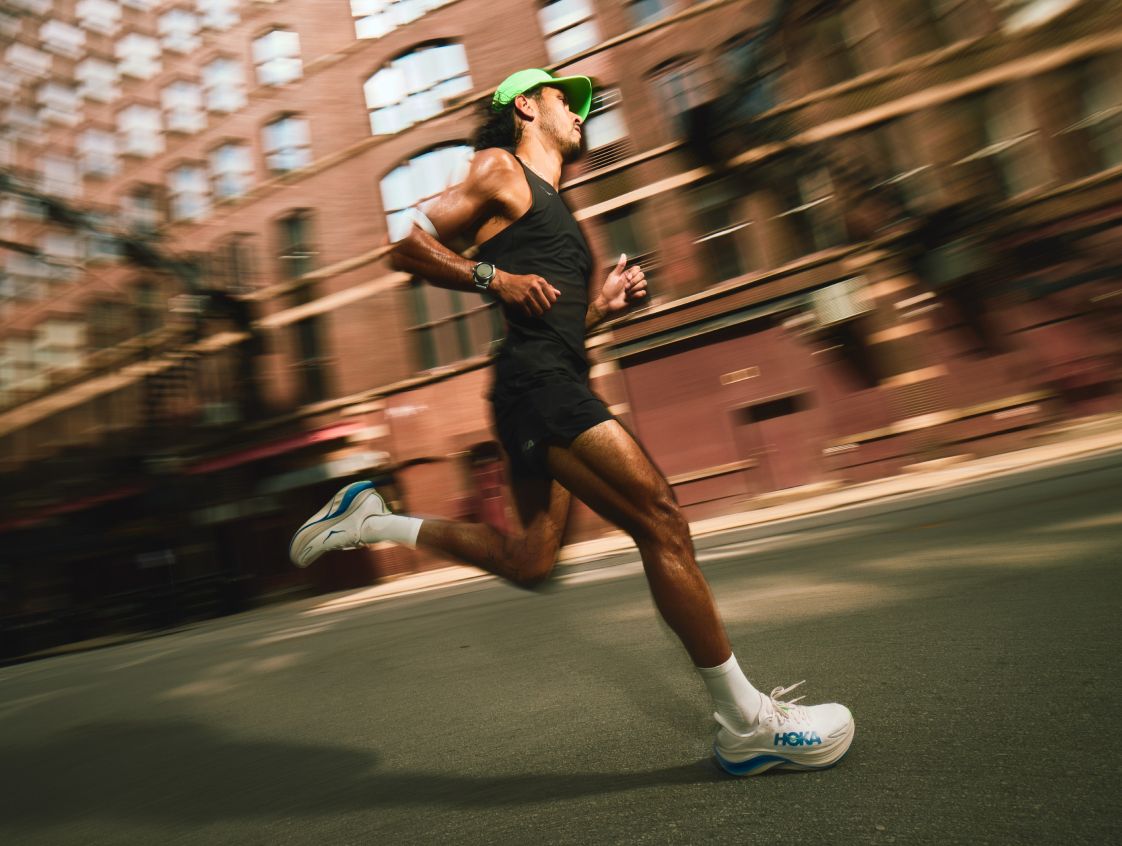 Vue latérale d’un coureur dans une rue de la ville montrant des chaussures de course HOKA blanches et bleues.