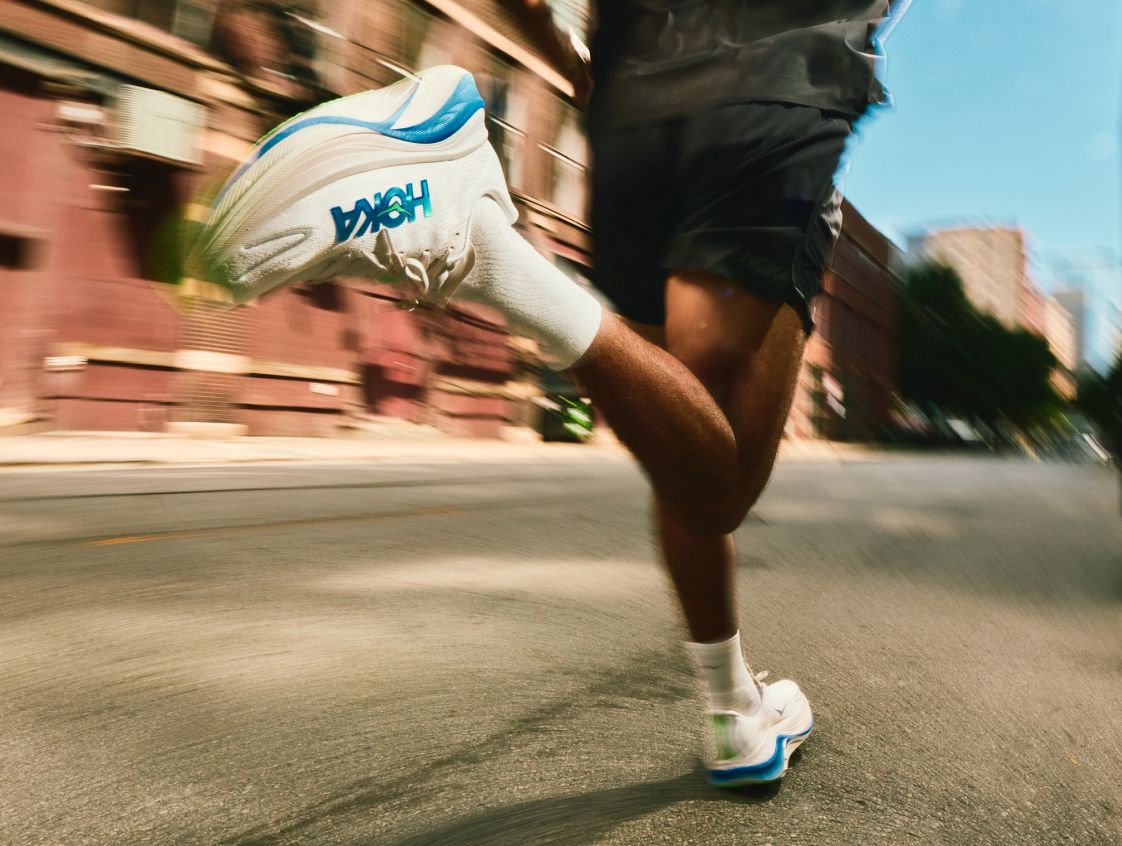 Photo d’action en pleine foulée se concentrant sur des baskets de course HOKA à semelles épaisses, blanches et bleues, avec une marque visible.