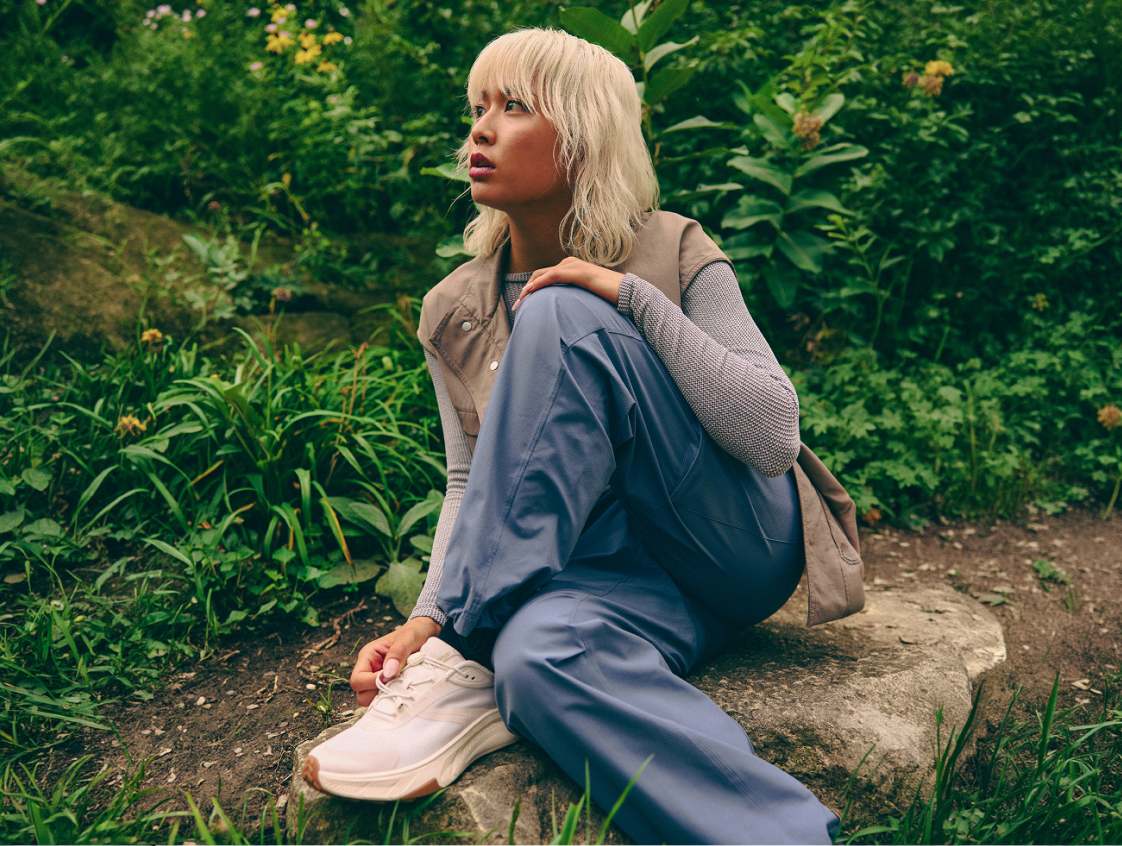 Une personne en gilet beige et pantalon bleu est assise sur un rocher parmi la verdure, portant des baskets blanches HOKA.