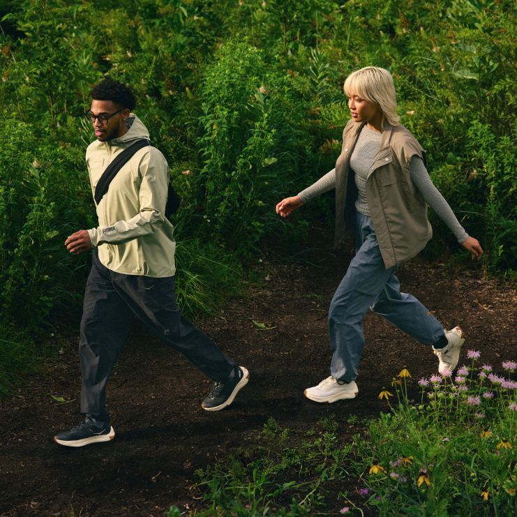 Deux personnes en tenue de plein air décontractée et baskets HOKA marchent le long d’un chemin de terre à travers des plantes verdoyantes et des fleurs sauvages.