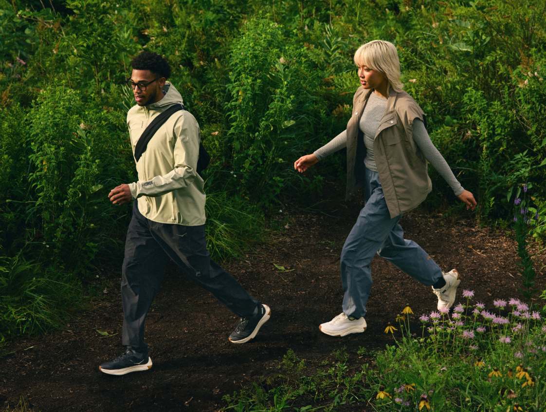 Deux personnes en tenue de plein air décontractée et baskets HOKA marchent le long d’un chemin de terre à travers des plantes verdoyantes et des fleurs sauvages.