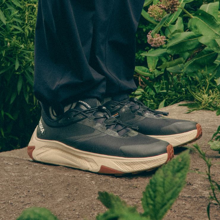 Gros plan des pieds d’une personne portant des baskets noires HOKA Transport avec semelles crème, debout sur un rocher entouré de feuillage vert.