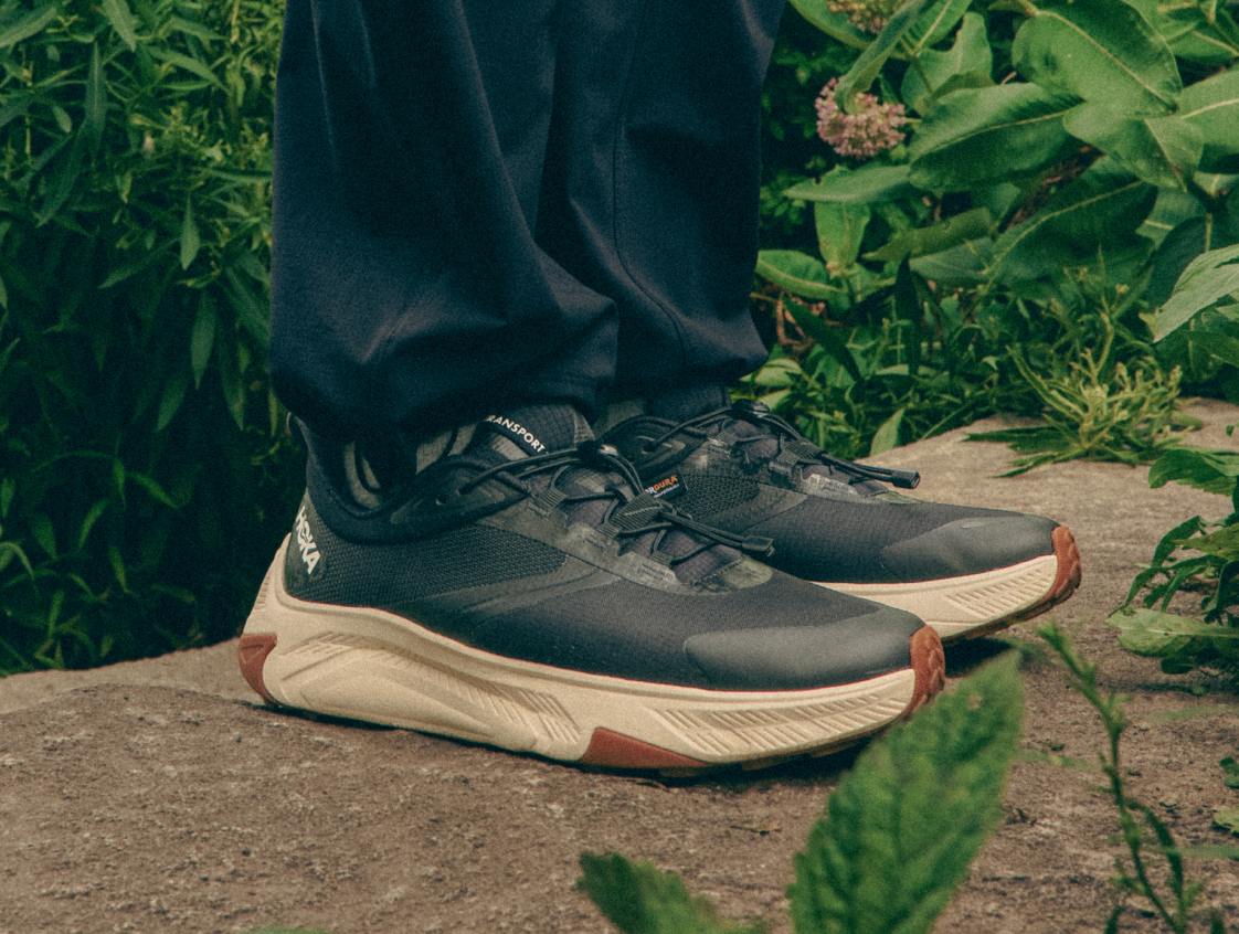 Gros plan des pieds d’une personne portant des baskets noires HOKA Transport avec semelles crème, debout sur un rocher entouré de feuillage vert.