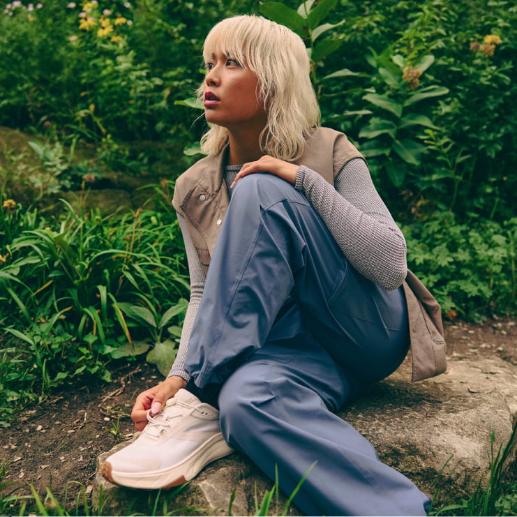 Une personne en gilet beige et pantalon bleu est assise sur un rocher parmi la verdure, portant des baskets blanches HOKA.