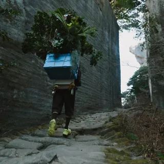 A person climbing a rocky hill while carrying a backpack with supplies and wearing 'HOKA Mafate Three2 Grid JP'